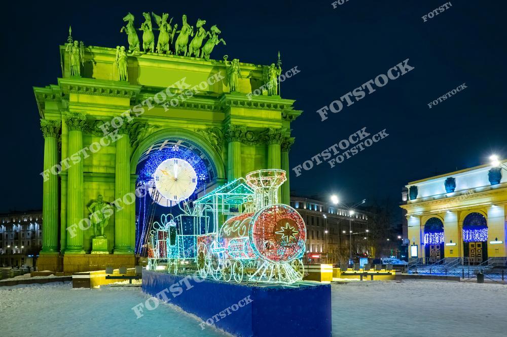 На фотографии изображены Нарвские триумфальные ворота в Санкт-Петербурге, украшенные праздничной иллюминацией. Перед аркой установлен светящийся поезд, символизирующий новогодние и рождественские праздники. Снежный покров и огни ночи подчеркивают торжественную атмосферу зимнего города. Эта композиция отражает историческое величие Петербурга в сочетании с праздничной красотой.